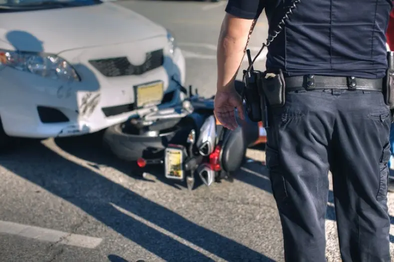 Un Oficial De Policía De Espaldas Observa La Escena De Un Accidente De Tráfico Con Un Automóvil Blanco Y Una Motocicleta En El Suelo.