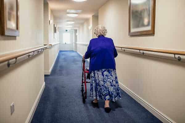 An elderly woman walks down a hallway at a nursing home using a walker.