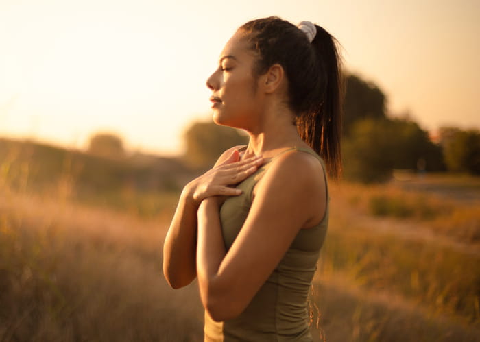 A woman breathing deeply with her hands crossed against her chest as she focuses on self-care in a field.