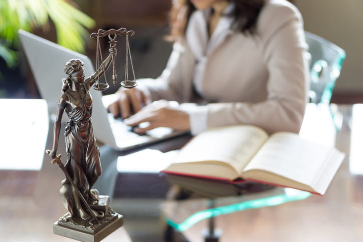 a lawyer is in the background, out of focus, while working on her laptop. In focus is a blindfolded statue holding the scales of justice in one hand and a sword in another.