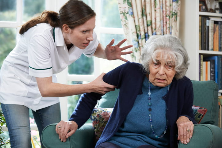 An image portraying nursing home abuse as an aid has her hand wrapped around an elderly woman's arm as she yells at her. The elderly woman looks scared as she tries to get out of her chair.