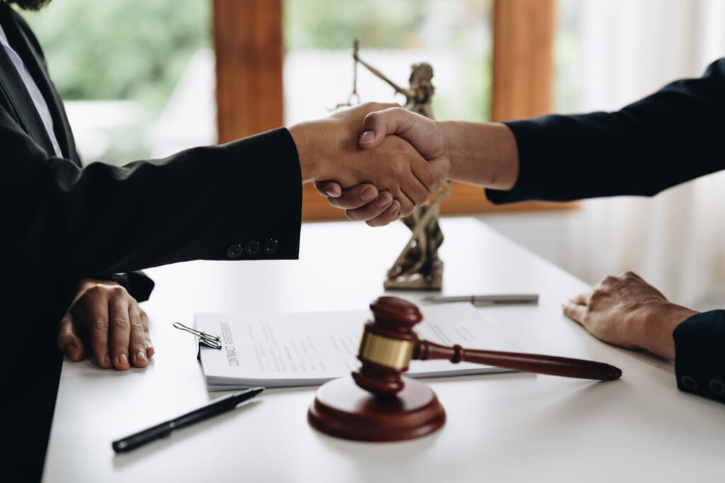 Birth injury lawyers shaking hands with a desk between them. On the desk is paperwork, a Lady Justice statue, and a gavel
