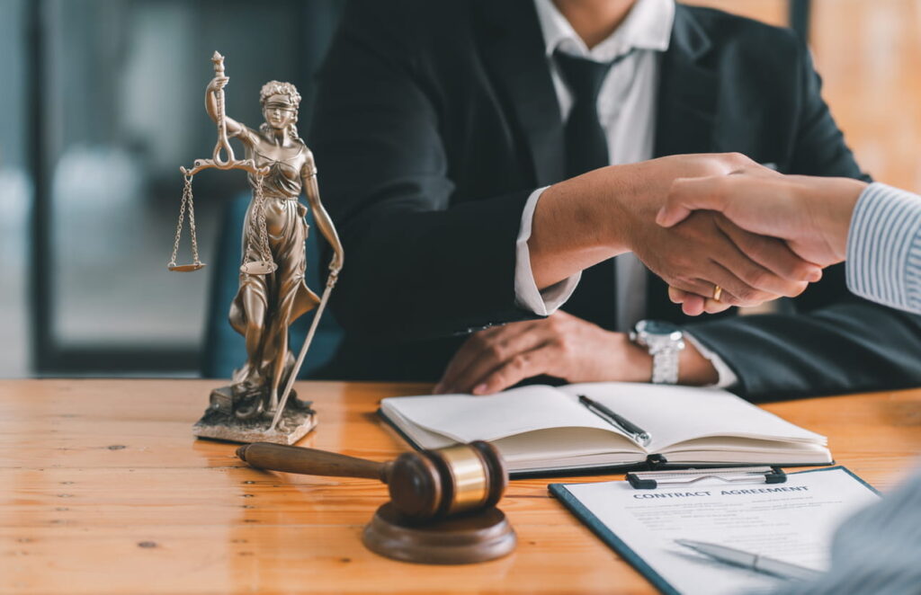 A pedestrian accident attorney shaking their clients hand. On their desk is paperwork, a Lady Justice statue, and a gavel.