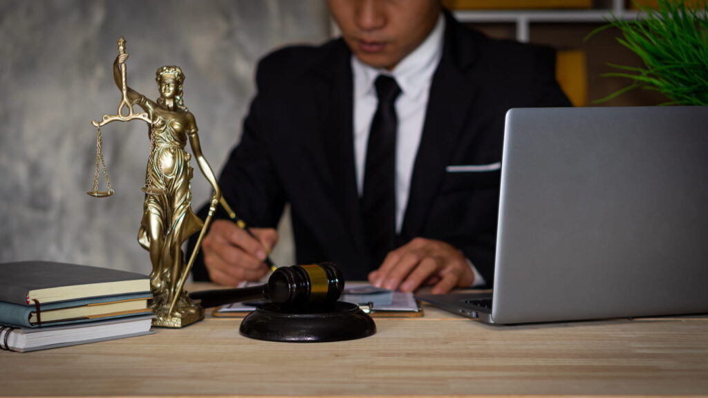 A car accident lawyer works on a case at his desk. In front of him is paperwork, an open laptop, a pile of books, the Lady Justice statue and a gavel.