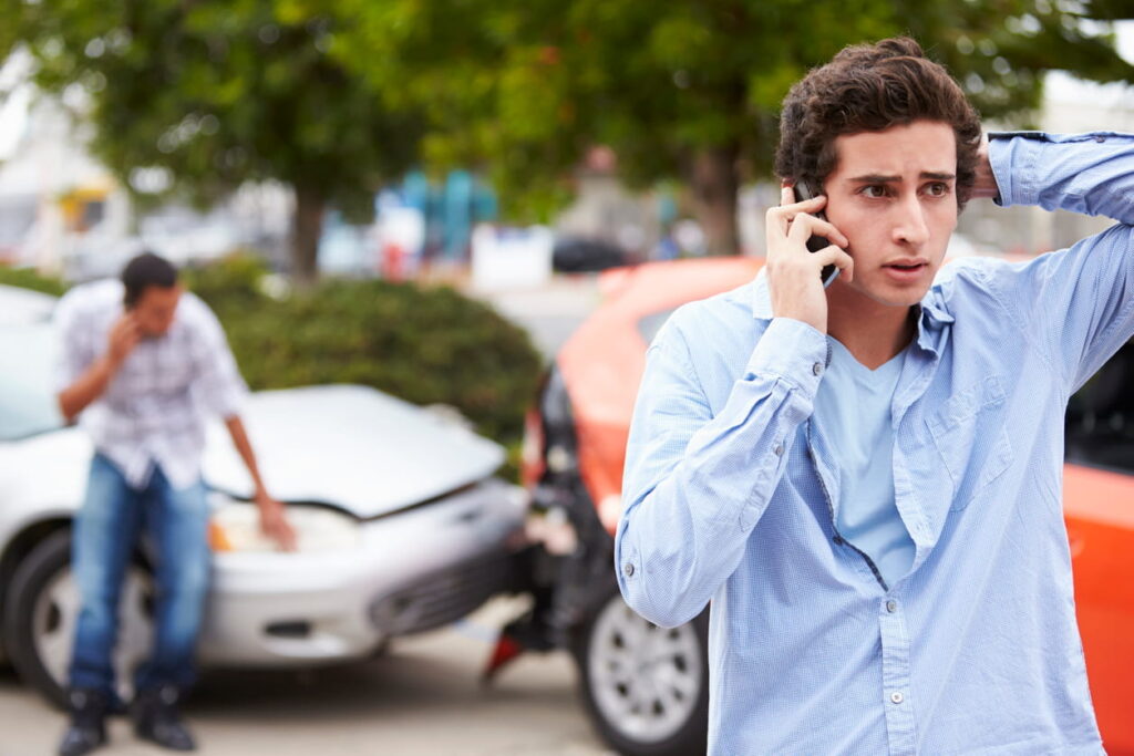 A concerned young man on tthe phone after suspecting a car accident scam. In the background is a minor car accident, with another man also on his phone.