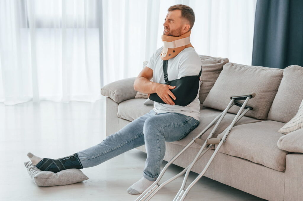 A man suffering from personal injuries. He's in a neck brace, his arm is in a sling, and he has a brace on his leg that's resting on a pillow. Next to him on the couch are crutches.