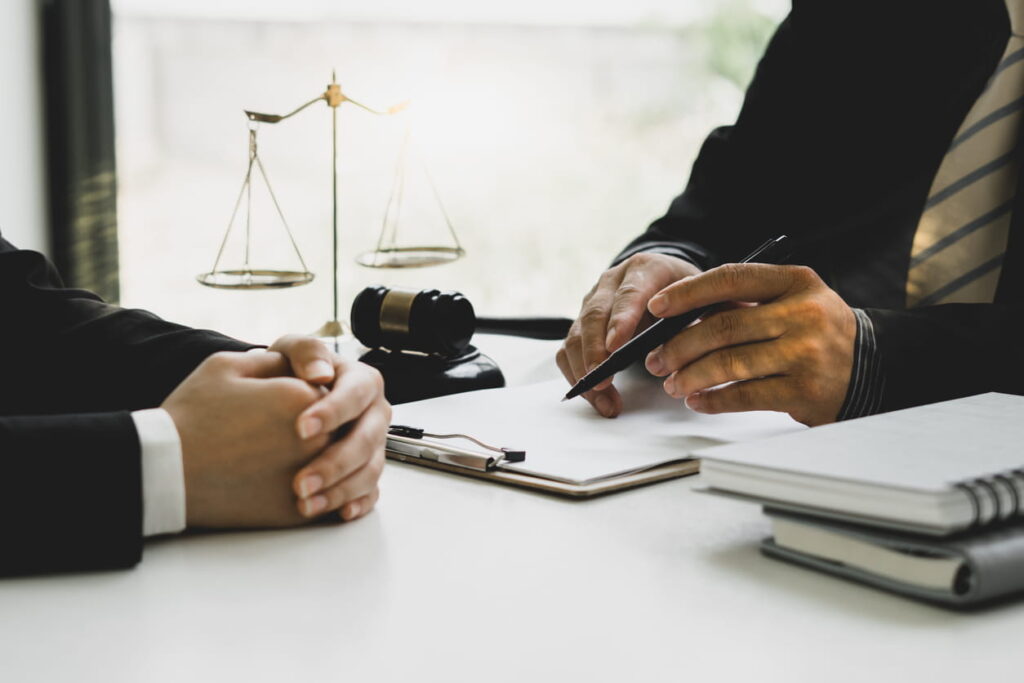 A personal injury lawyer is working with a client at his desk. In front of him is paperwork, a stack of notebooks, the scales of justice and a gavel.