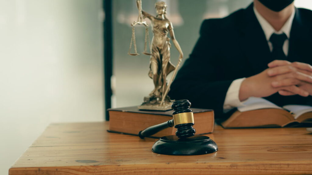 An employment law attorney sits at his desk with his hands together next to a stack of books with a Lady Justice statue on top, and a gavel laying next to it.