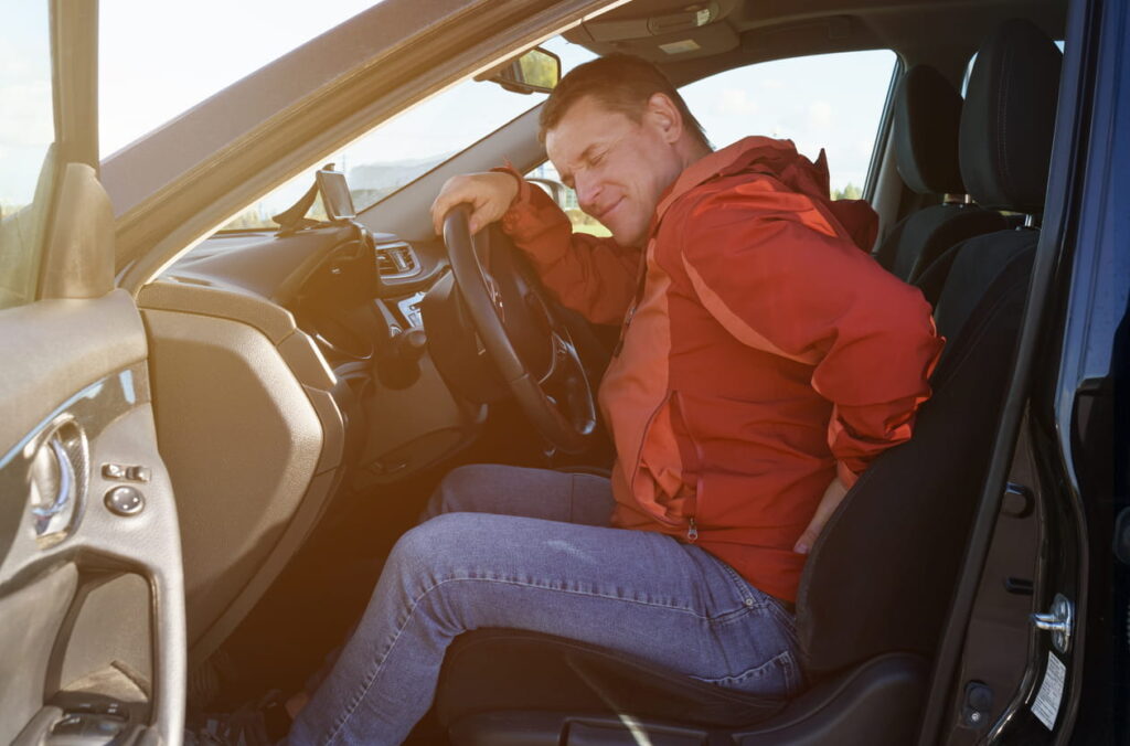 A man holding his lower back in pain, while sitting behind the wheel of his car after an accident.