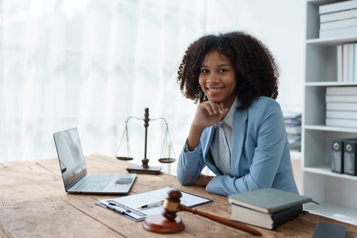 A personal injury attorney resting her chin on her hand at her desk, smiling for the camera. On her desk is an open laptop, paperwork, the scales of justice, a gavel, and a stack of books. 