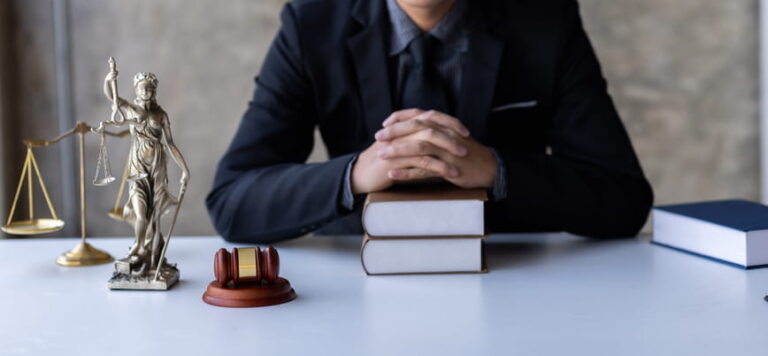 A wrongful death attorney sits with his hands folded on a stack of books. Next to him is the scales of justice, a Lady Justice statue, a gavel and another book. 