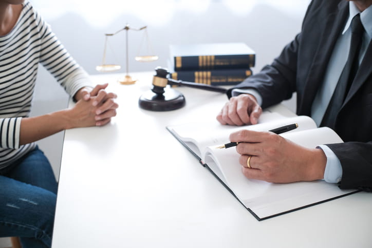 A personal injury attorney assisting a client on their case. Between them is an open notebook, a gavel, the scales of justice and a stack of books. 