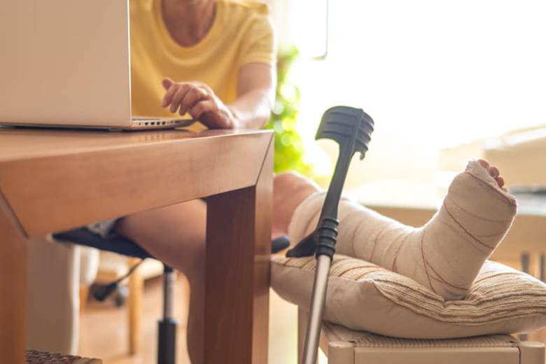 A woman sits at the table with her laptop as her injured leg is propped up on a stool.