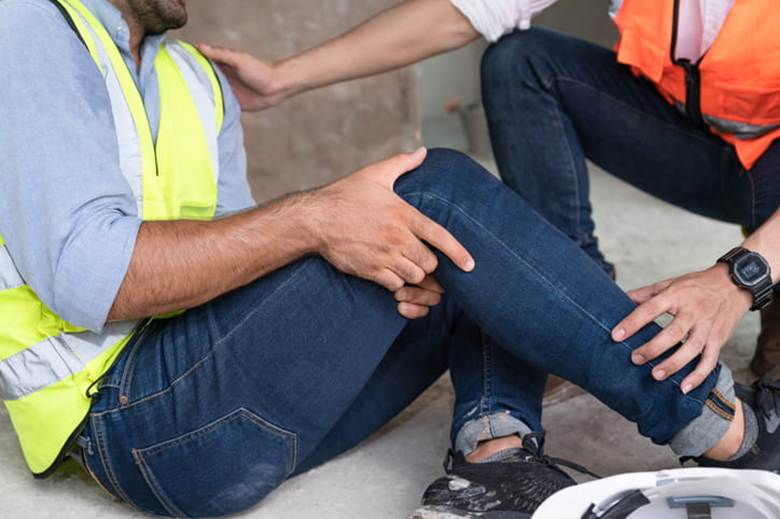 An injured worker grabs his knee in pain as a coworker assists him.